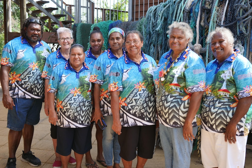   One man and seven women all dressed in colourful blue shirts, standing in front of ropes.