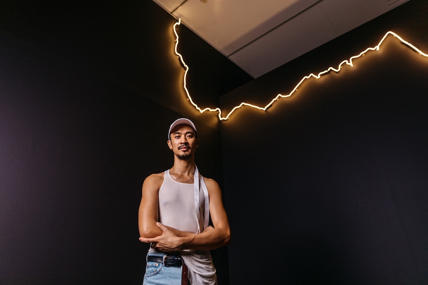 Tanned man with dark hair and trim beard wears white singlet and cap and stands with folded arms in front of light installation