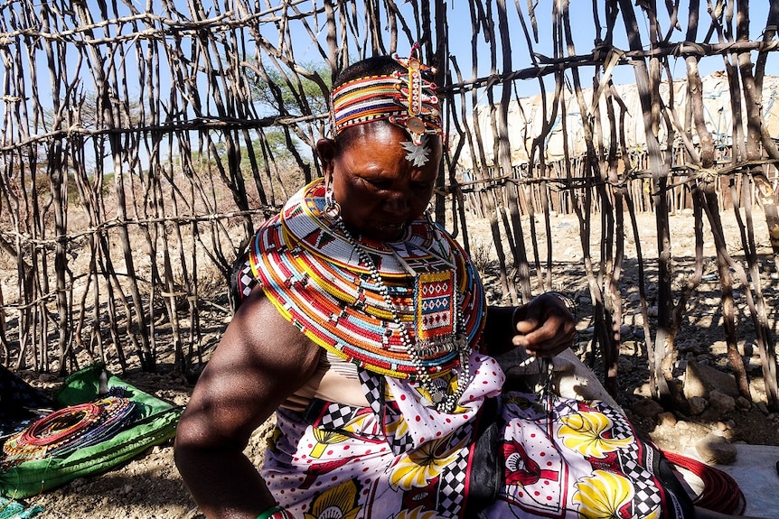 A woman wearing colourful beadwork sits in a remote hut during the day.