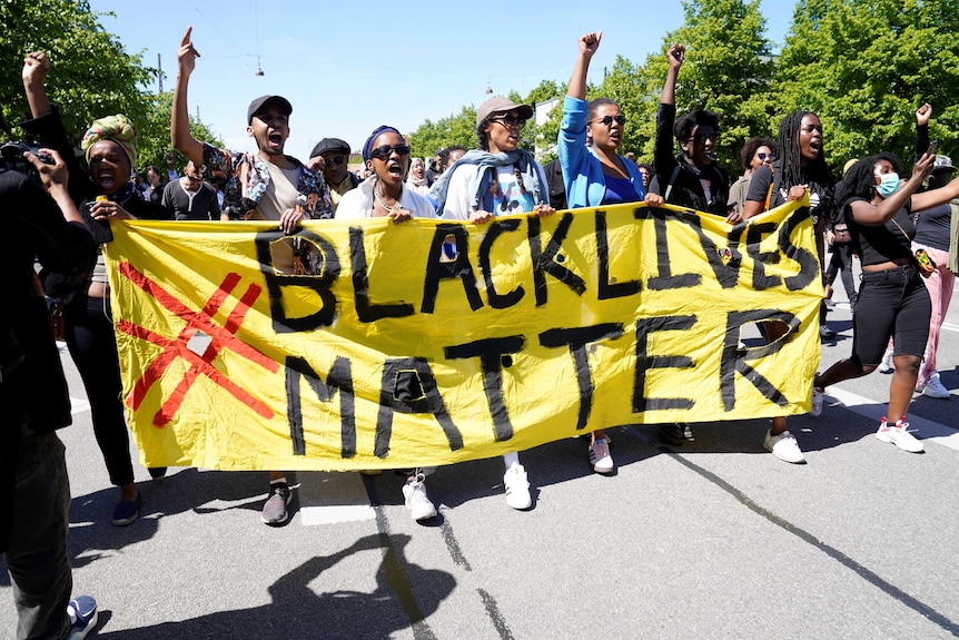 On a bright blue day, you view a line of people of African descent holding a yellow banner that reads 'Black Lives Matter'.