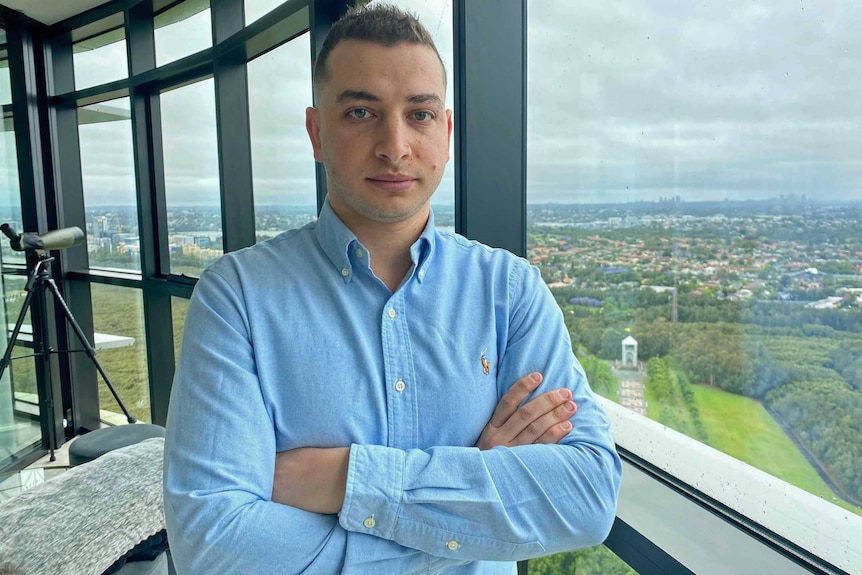 Man stands with arms crossed near windows.