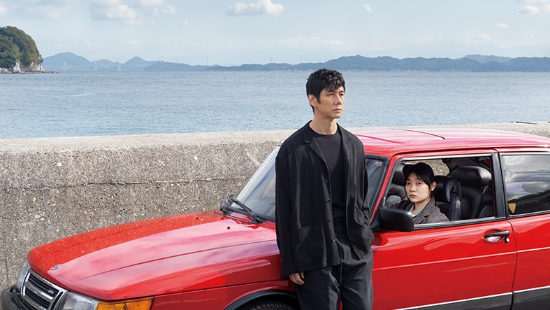 Japanese man wears all black and leans against red sedan looking wistful while Japanese woman sits in driver's seat looking up.