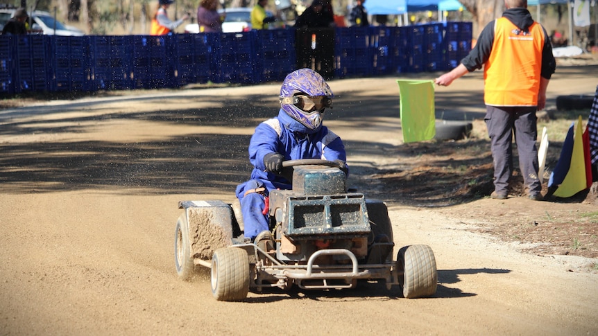 A man in a blue race suit rides a lawn mower around a track kicking up dirt. 