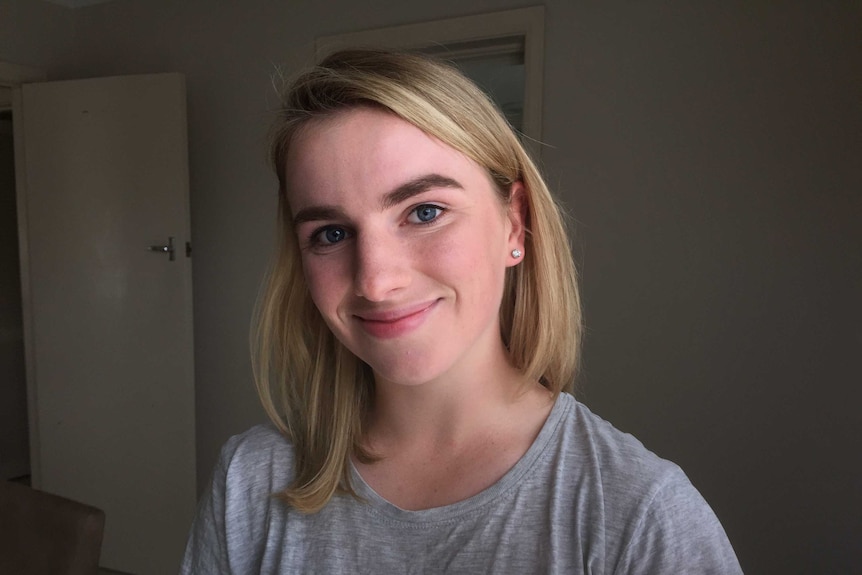 Adeline Collins smiles at the camera. She is wearing a plain grey T-shirt and standing in front of an open door.