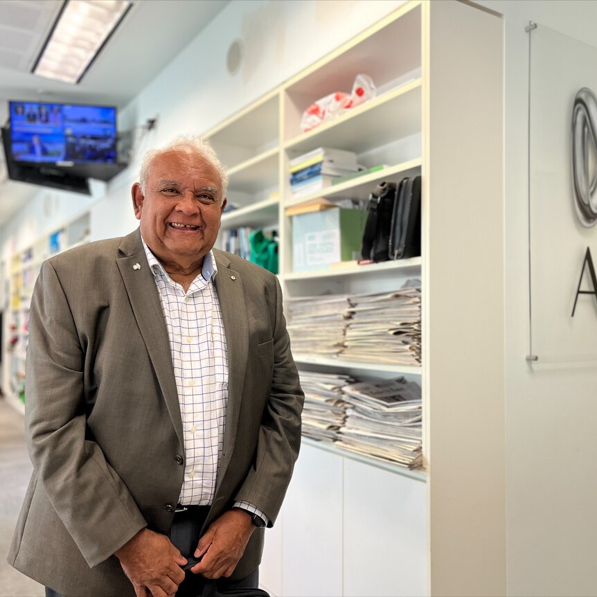 Tom Calma smiles brightly at the camera in front of an abc sign, he wears a suit jacket and a striped shirt