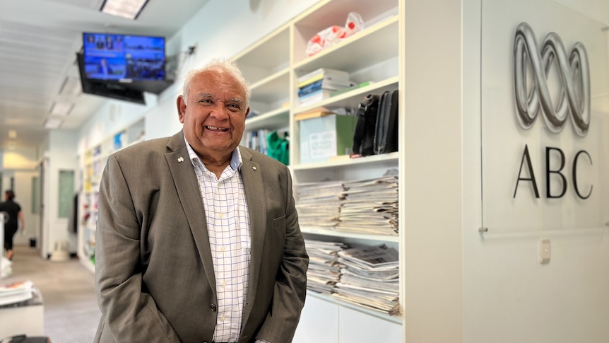 Tom Calma smiles brightly at the camera in front of an abc sign, he wears a suit jacket and a striped shirt