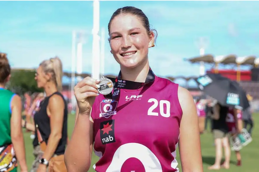 A young AFLW player in a maroon uniform holding up a medal she's wearing around her neck.