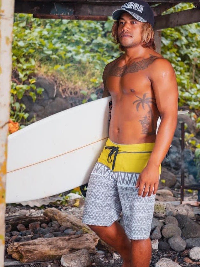 A Samoan man with his shirt off, tatoos and boardshorts holding his board, blonde surfie hair and a cap