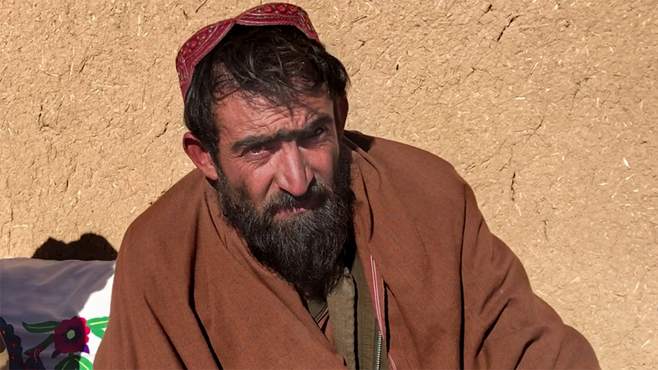 Sayed Jan, beared with black hair wearing a red cap, sits in front of a light brown wall outside.