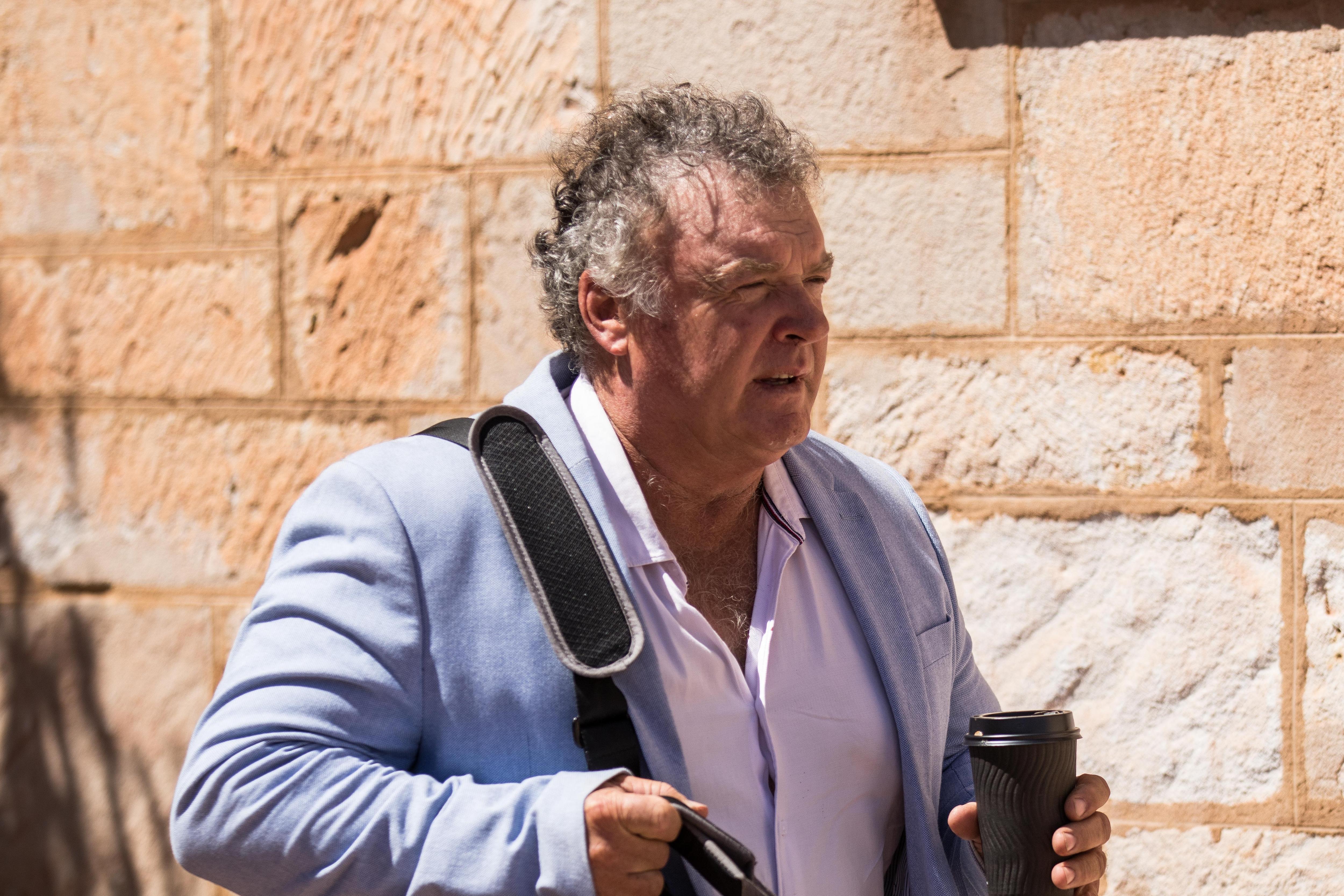 A man in a suit jacket arrives at a courthouse carrying a bag and coffee cup.  
