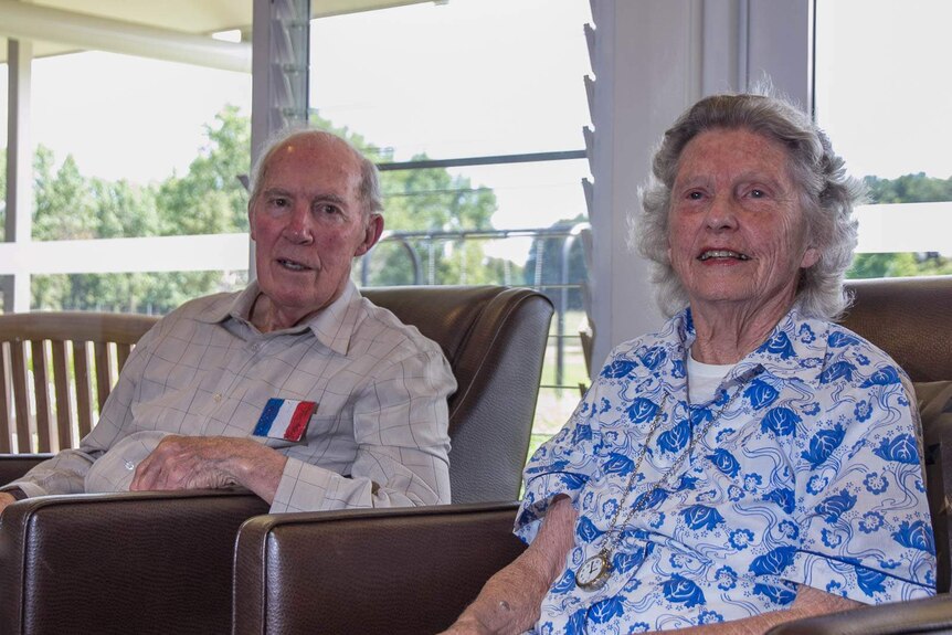 An elderly man and woman in arm chairs at a nursing home in front of a window with sun coming in