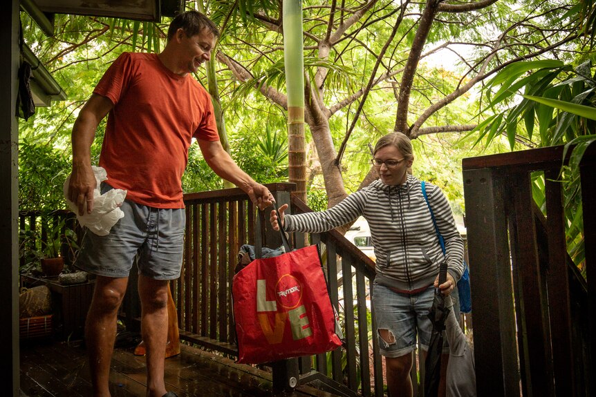 a man hands a red bag full of clothing to a woman who is standing on the steps of his verandah with tress in the background