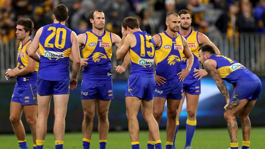 A group of West Coast Eagles players stand on Perth Stadium looking dejected after losing a game.