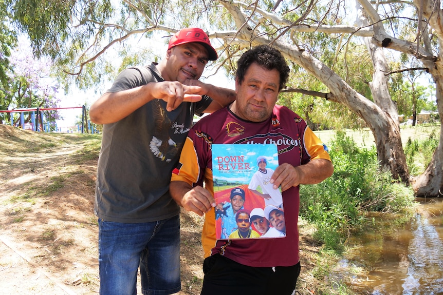Two men standing in front of the river with The Wilcannia Mob book.