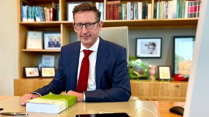 Stephen Jones sits at a desk. He wears a suit. A large bookcase is behind him.