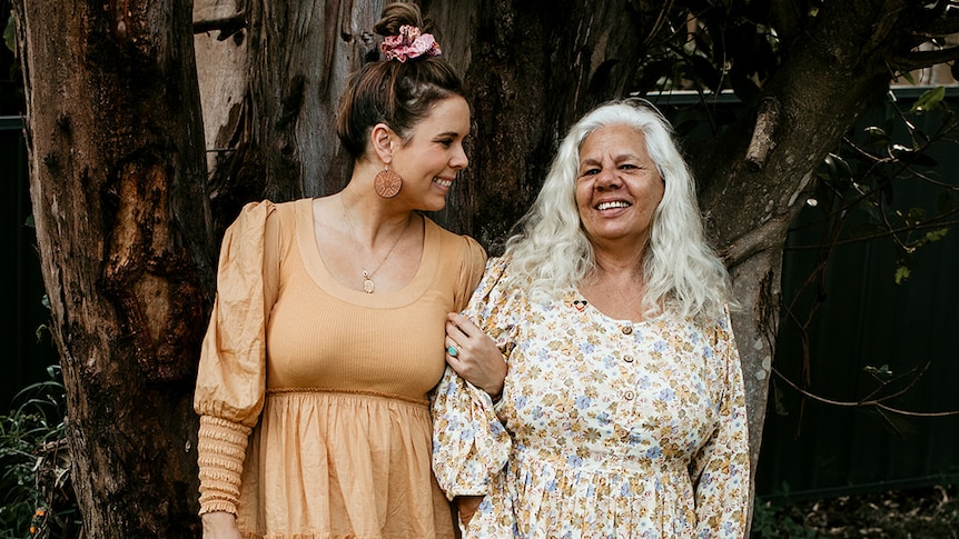 A young Aboriginal woman stands with her arm interlocked with her mum, outside, both smiling.