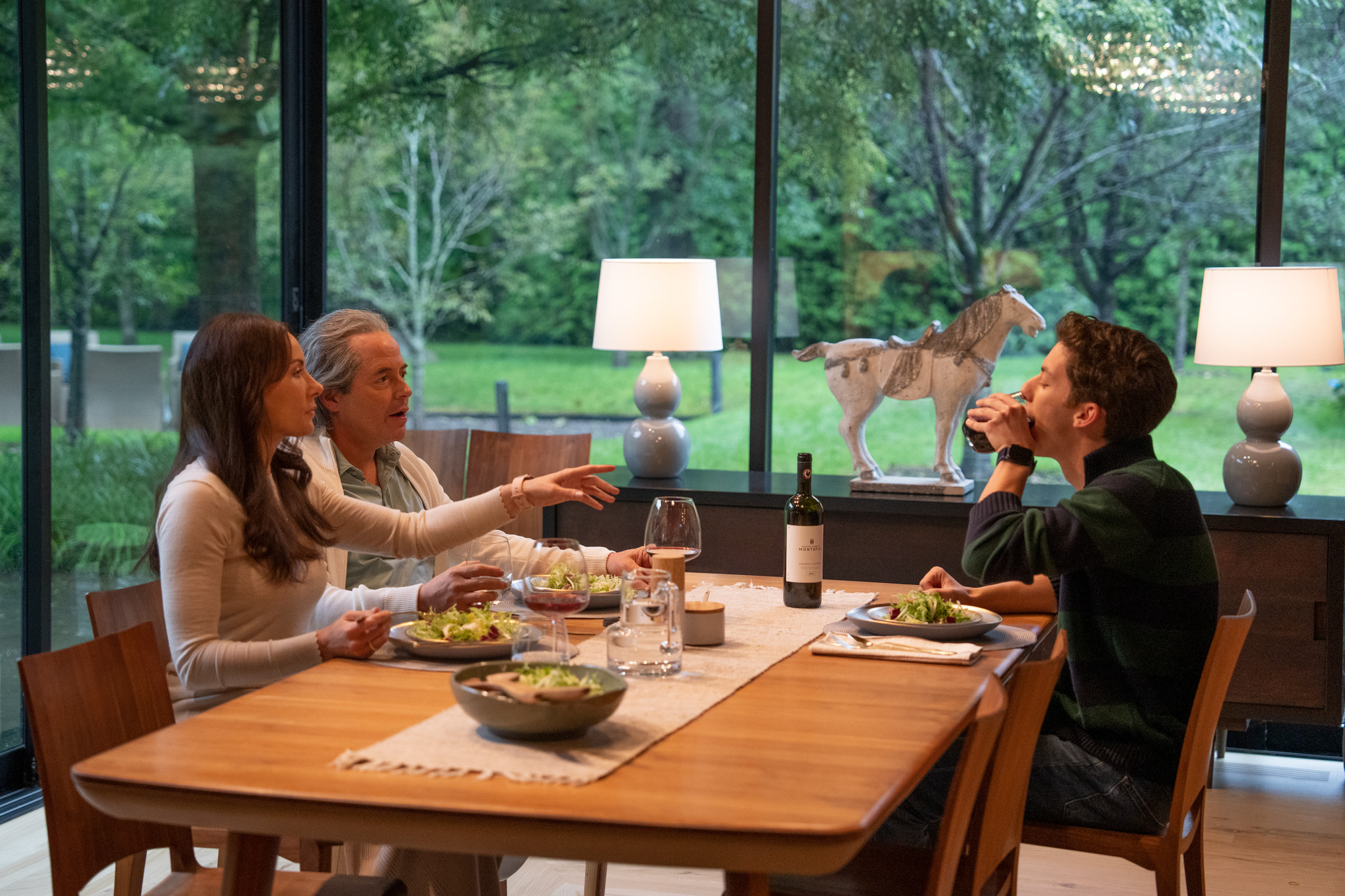 A middle-aged white woman and man and a white teen boy sit at an elegantly set dinner table overlooking a garden.