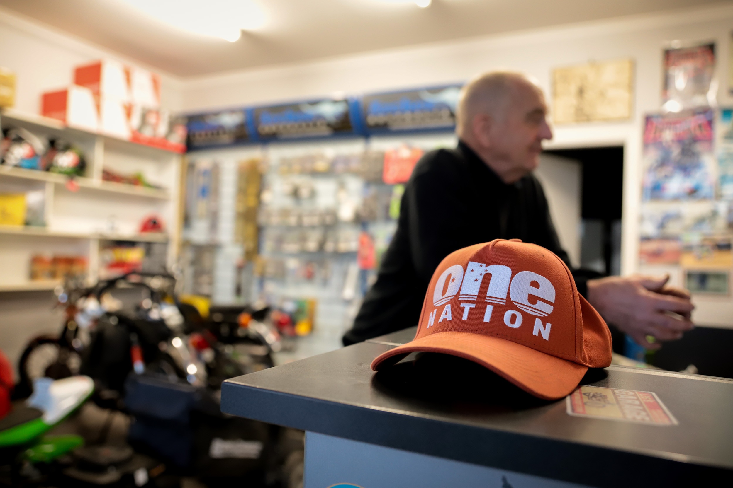 Orange hat with One Nation text sits on bench with man standing behind inside shop 