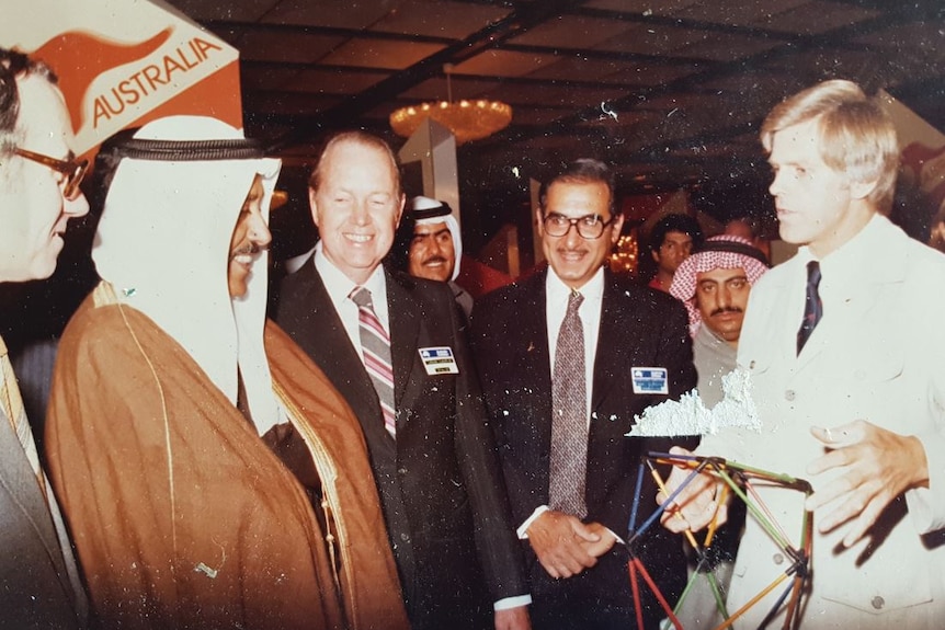 A grainy photo of man with white suit and blonde hair standing with group of men, wearing either suits or headscarves.