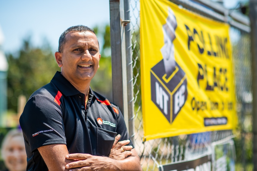 A man wearing a black and red polo shirt stands next to a wire fence with 'polling place' affixed to a banner.
