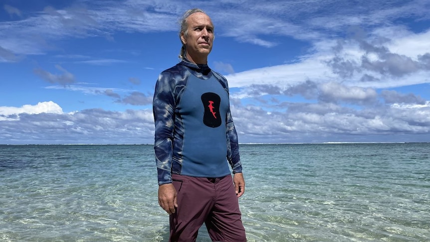 A man with grey hair wearing a blue multicoloured sleeved rash vest stands in crystal blue water on a blue sky day