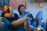 A couple are laughing and sitting on a couch watching tv with a bottle of wine and glasses in front of them