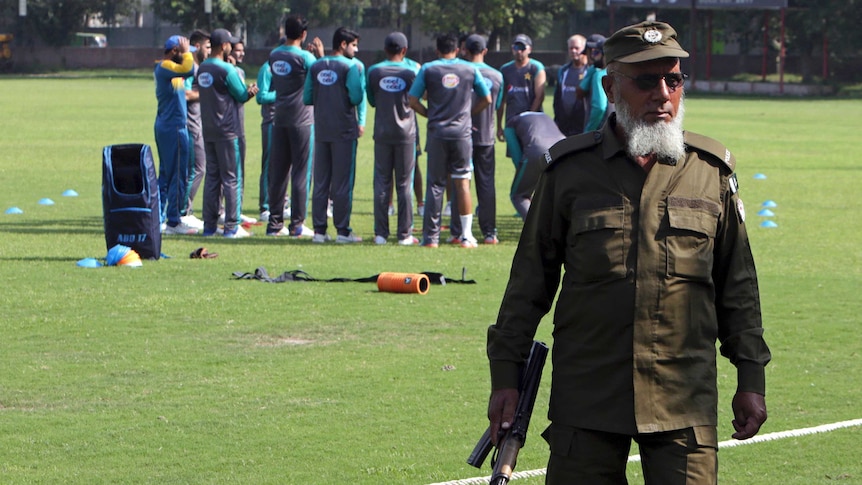 A police officer in the foreground holds an AK-47 while Pakistan's cricket team trains behind him.