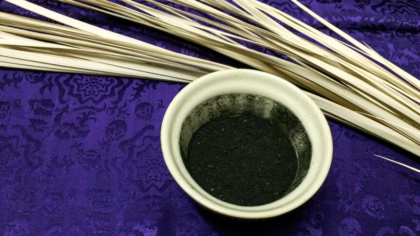 Close up photo of a bowl of fine grey ash and a dried palm front resting on a purple cloth.