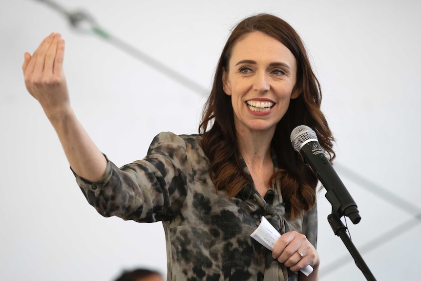 A woman with black hair wearing a green and black top gestures with her hands while in front of a microphone