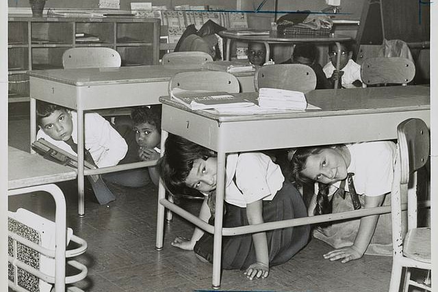 A black and white image of children crouching under their desks at school.
