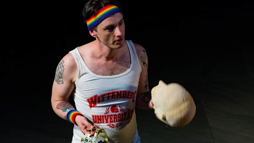 Dickie Beau in a singlet and rainbow headband holds a crown in one hand and a mannequin head in the other.
