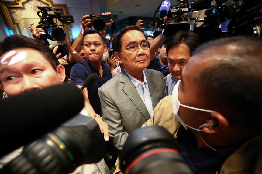 A Thai man in grey suit with no tie and thin-rimmed glasses stands unsmiling in crowd of photographers.