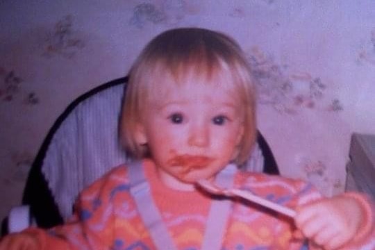 Blonde haired baby sitting propped up in high-chair smiles, food all over her face, eating from a spoon.