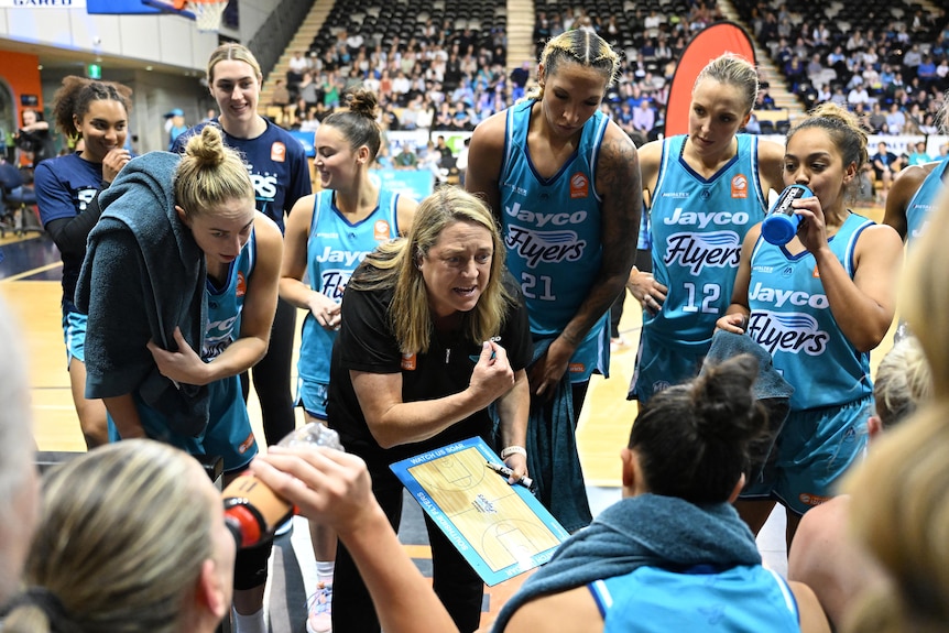 A WNBL basketball coach stares down one of her players while the rest of the players gather round during a team talk.