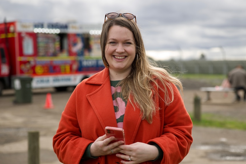 a woman is smiling at the camera.  She is holding a phone and wearing a red jacket.