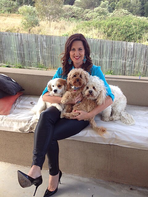 A woman sits on a bench inside cuddling three small white dogs.