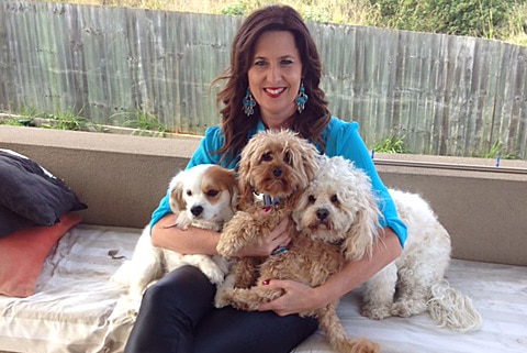 A woman sits on a bench inside cuddling three small white dogs.