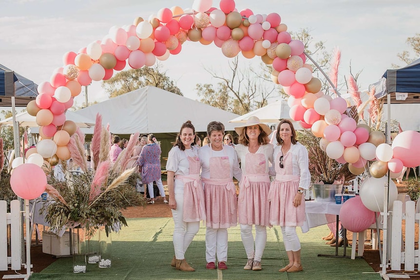 Four women wearing matching aprons stand in front of a balloon archway