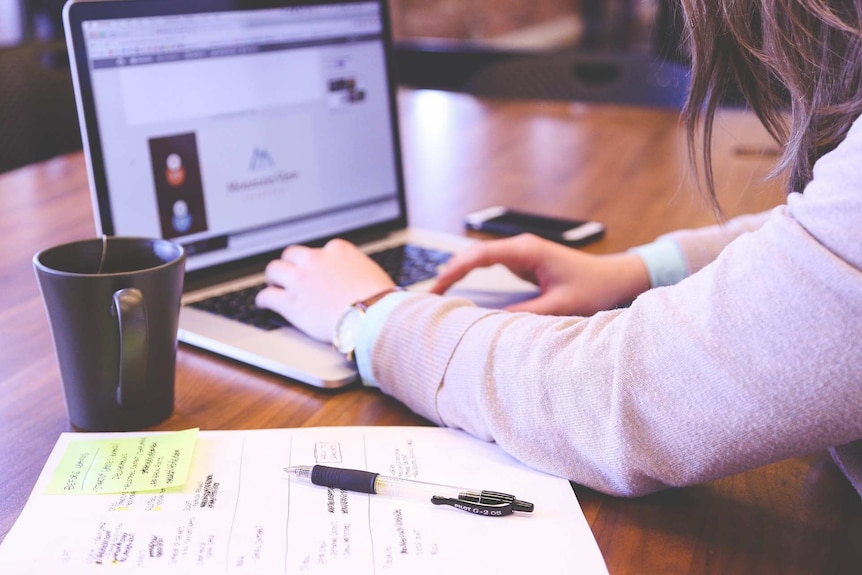 A woman works on a laptop. A cup of tea and some documents sit near her elbow.