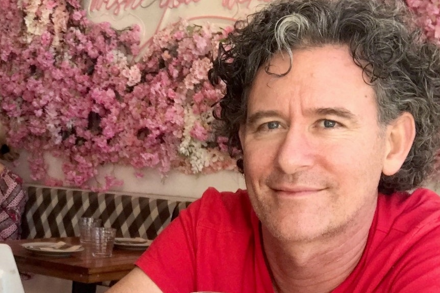 A middle-aged man in a red t-shirt sits in a cafe with a floral feature on the wall.