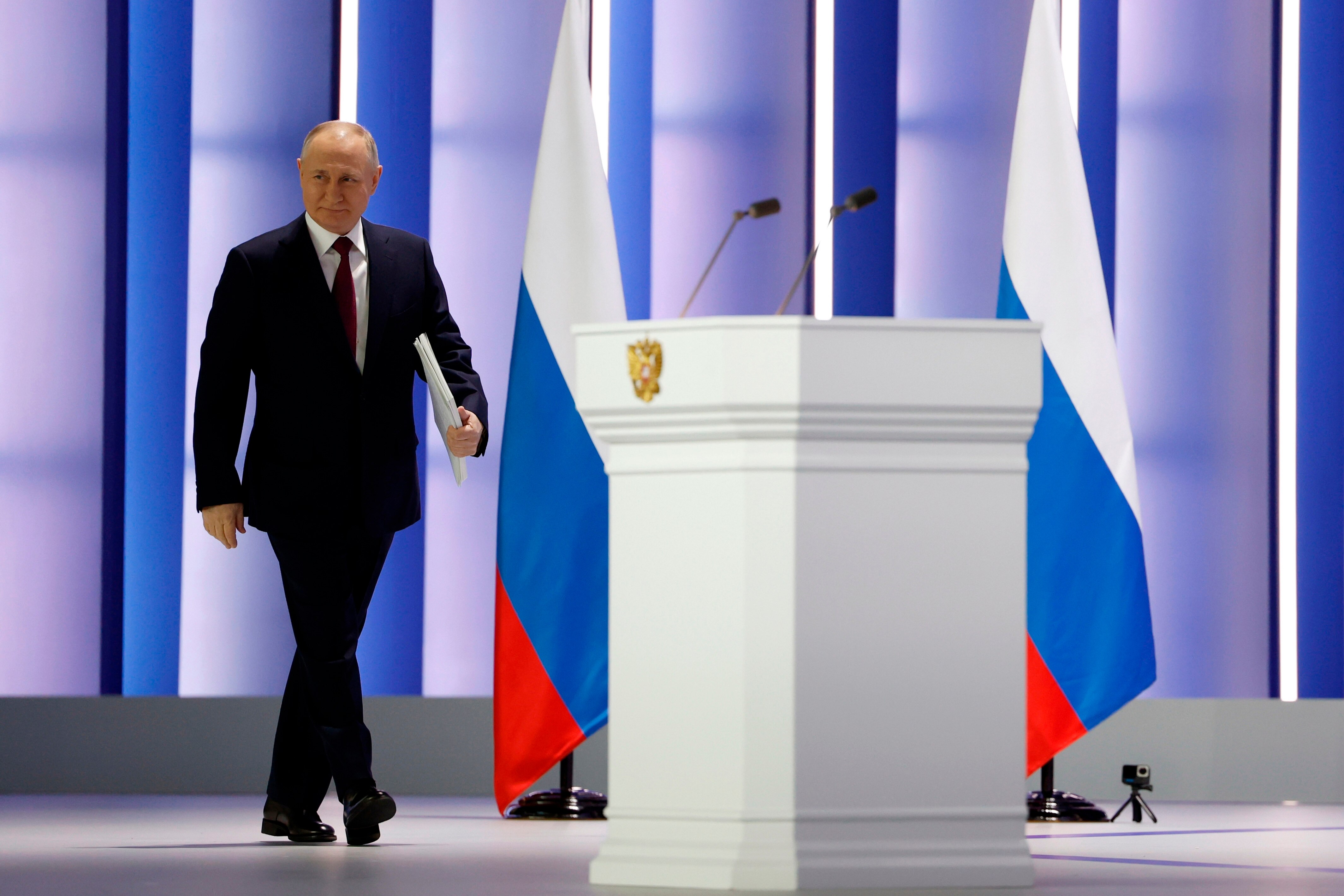 Vladimir Putin approaches a podium on a well lit stage with Russian flags visible in background.