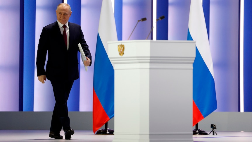 Vladimir Putin approaches a podium on a well lit stage with Russian flags visible in background