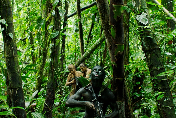 Teen boy painted in black paint looks up while holding rifle and leads a teen girl and boy painted in mud through thick jungle.