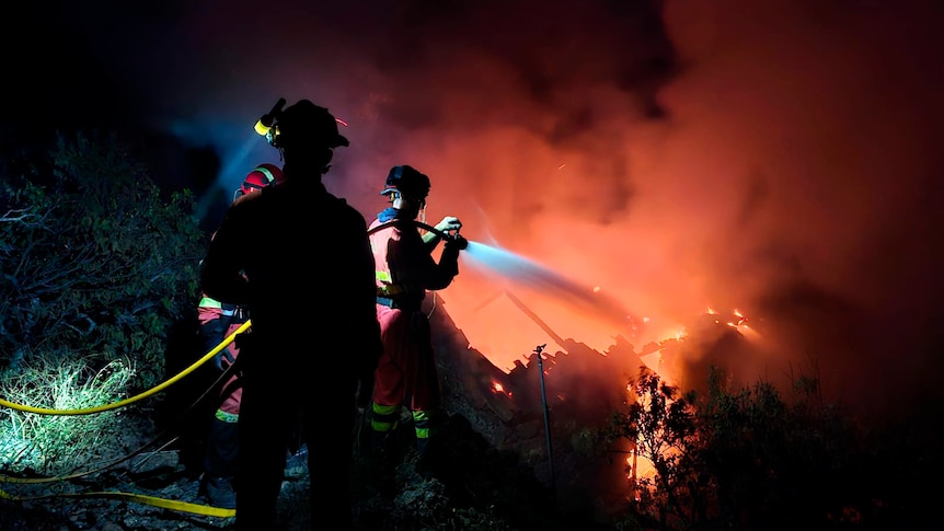 Fire lights up the sky with an orange and red glow as firefighters can be seen holding hoses in the dark.