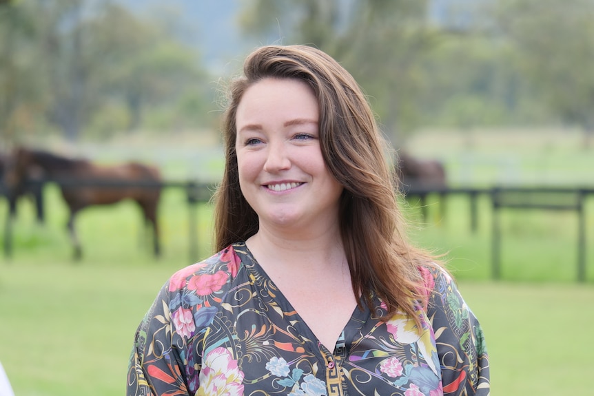 A smiling woman standing in a paddock with horses in the background