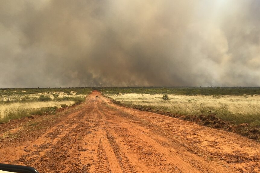Heavy grey smoke from a bushfire over a  wide, red dirt road through scrub.