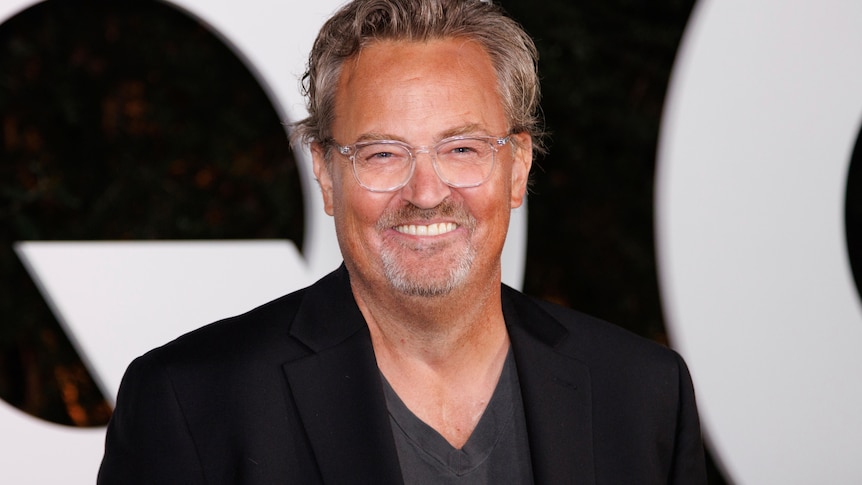A close up of a smiling Matthew Perry, wearing clear plastic-rimmed glasses, a black suit jacked and a grey v-neck shirt. 