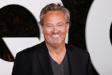 A close up of a smiling Matthew Perry, wearing clear plastic-rimmed glasses, a black suit jacked and a grey v-neck shirt. 