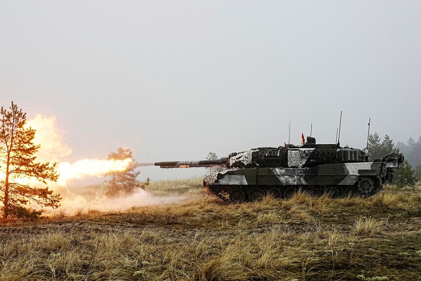 A dark-green and white patterned tank fires an explosive round on a grassy field.