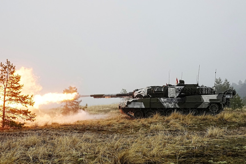 A dark-green and white patterned tank fires an explosive round on a grassy field.